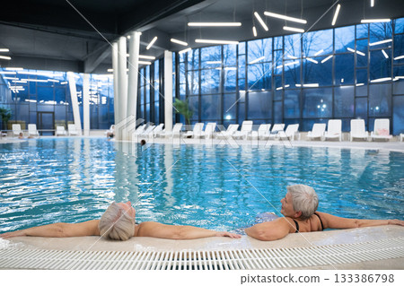 Two senior women enjoying relaxation while leaning on the edge of a modern indoor swimming pool in the evening. Concept of wellness, healthy lifestyle, spa, leisure, and friendship for older adults. Two senior women enjoying relaxation while leaning on the edge of a modern indoor swimming pool in the evening. Concept of wellness, healthy lifestyle, spa, leisure, and friendship for older adults. 133386798