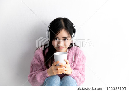 A young woman sitting and thinking while listening to music on headphones with her back to a white wall 133388105