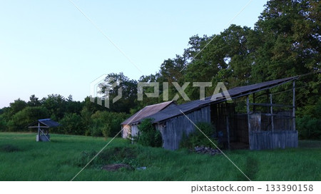 An old dilapidated barn for storing hay and a retro well on a field with lush tall grass near the forest on a summer evening at dusk, abandoned village buildings on an evening summer meadow An old dilapidated barn for storing hay and a retro well on a field with lush tall grass near the forest on a summer evening at dusk, abandoned village buildings on an evening summer meadow 133390158