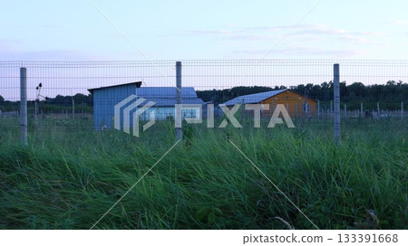 Outbuildings on farmland behind a chain-link fence on an unmown lawn, rural landscape with a house, barn and fence in the evening summer twilight, residential and working farmland Outbuildings on farmland behind a chain-link fence on an unmown lawn, rural landscape with a house, barn and fence in the evening summer twilight, residential and working farmland 133391668
