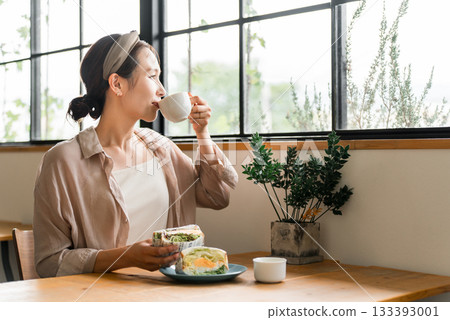 一位亞洲女性在咖啡館吃三明治、喝咖啡，可能是午餐、早午餐或早餐。 133393001