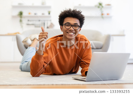 A young Black man with glasses smiles while giving a thumbs up, using a laptop on the floor. A young Black man with glasses smiles while giving a thumbs up, using a laptop on the floor. 133393255