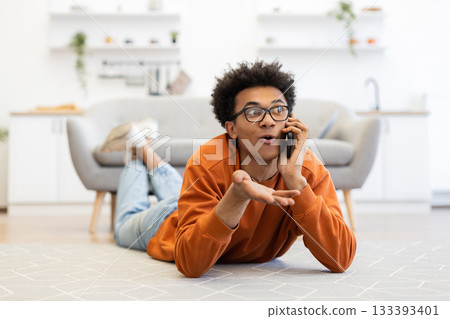 A young man with glasses lies on the floor, talking on his phone with an expressive gesture in a modern living room. A young man with glasses lies on the floor, talking on his phone with an expressive gesture in a modern living room. 133393401