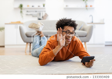 A young man with glasses lies on the floor, waving at his phone during a video call in a cozy living room. A young man with glasses lies on the floor, waving at his phone during a video call in a cozy living room. 133393409