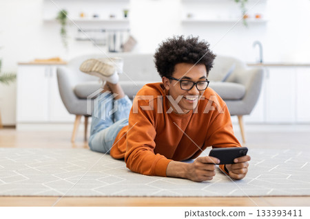 A young man lies on the floor, smiling while looking at his phone in a cozy living room setting. 133393411