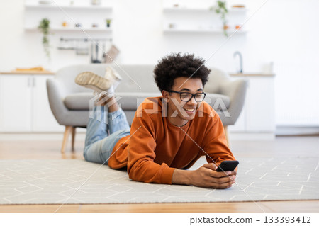 A young man with glasses lies on the floor, smiling while using his smartphone in a cozy living room. A young man with glasses lies on the floor, smiling while using his smartphone in a cozy living room. 133393412