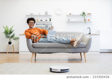 A young man relaxes on a couch with a laptop and remote control while a robot vacuum cleans the floor. A young man relaxes on a couch with a laptop and remote control while a robot vacuum cleans the floor. 133393416
