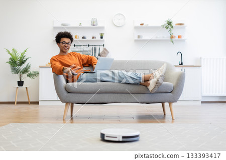 A young man relaxes on a sofa with a laptop and remote control while a robot vacuum cleans the floor. A young man relaxes on a sofa with a laptop and remote control while a robot vacuum cleans the floor. 133393417