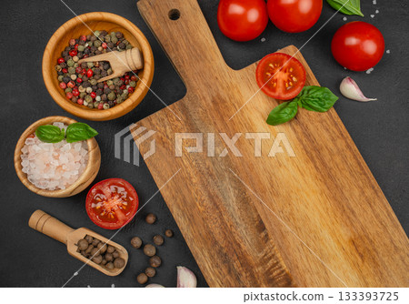 Fresh cooking ingredients tomatoes, basil, spices and garlic arranged around an empty wooden cutting board on dark stone background, top view copy space Fresh cooking ingredients tomatoes, basil, spices and garlic arranged around an empty wooden cutting board on dark stone background, top view copy space 133393725