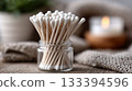 Cotton swabs in a glass jar on a wooden surface with a candle in the background 133394596