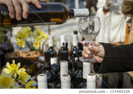 Close-up of a person's hand pouring white wine into a glass, many bottles of wine nearby 133400184