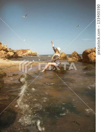 young blonde in a short dress and hat sits on a rock in the sea on a deserted beach. young blonde in a short dress and hat sits on a rock in the sea on a deserted beach. 133400390