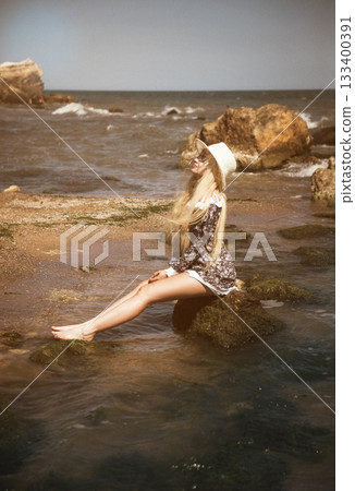 young blonde in short dress and hat sitting on rock in sea on beach 133400391