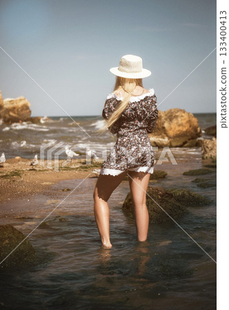 Blonde in a short summer dress and a light hat stands on the beach, with her back to the camera. 133400413