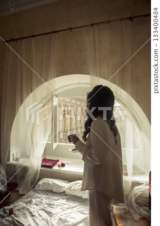 young brunette with long wavy hair stands in front of the window with a cup of coffee 133400484