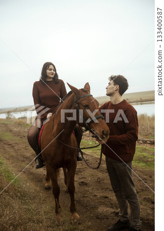 Young romantic couple horse riding, against the background of a field with autumn grass and a lake Young romantic couple horse riding, against the background of a field with autumn grass and a lake 133400587