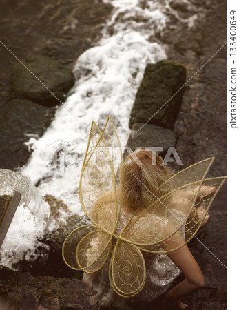 Cute blonde in fairy costume with wings and lace, transparent white dress sits in front of waterfall 133400649