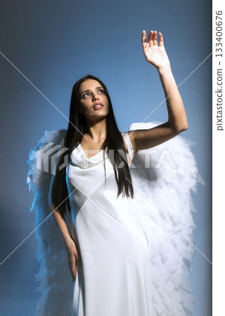studio photo of blonde in white long dress and angel wings posing on blue background in studio studio photo of blonde in white long dress and angel wings posing on blue background in studio 133400676