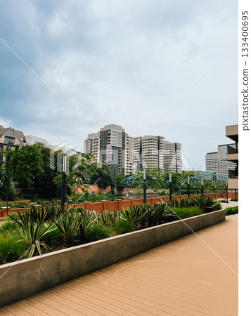 Urban park nestled between high rise residential buildings, featuring wooden walkway, verdant landscape, river passing under arched bridge, embodying contemporary metropolitan tranquility. Living area Urban park nestled between high rise residential buildings, featuring wooden walkway, verdant landscape, river passing under arched bridge, embodying contemporary metropolitan tranquility. Living area 133400695