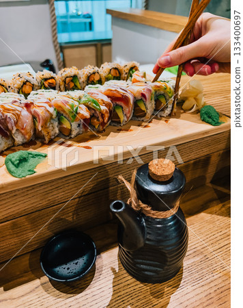 Wooden tray brimming with assortment of sushi rolls, including maki and uramaki, accompanied by wasabi, gari, and soy sauce, while customer skillfully picks up piece with chopsticks. Sushi rolls Wooden tray brimming with assortment of sushi rolls, including maki and uramaki, accompanied by wasabi, gari, and soy sauce, while customer skillfully picks up piece with chopsticks. Sushi rolls 133400697