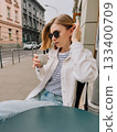 Young woman with sunglasses sitting at table, drinking coffee and adjusting her hair in street cafe in European city, enjoying quiet moment. Woman sipping coffee, while relaxing at outdoor cafe table 133400709