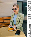Elegant woman wearing sunglasses, green jacket and blue turtleneck, sitting at wooden table outdoors, holding yellow cup of coffee and with smartphone next to it, enjoying break during sunny day 133400712