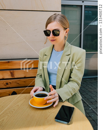 Elegant woman wearing sunglasses, green jacket and blue turtleneck, sitting at wooden table outdoors, holding yellow cup of coffee and with smartphone next to it, enjoying break during sunny day Elegant woman wearing sunglasses, green jacket and blue turtleneck, sitting at wooden table outdoors, holding yellow cup of coffee and with smartphone next to it, enjoying break during sunny day 133400712