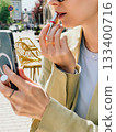 Professional woman checking smartphone while touching up red lipstick on urban sidewalk, reflecting modern workplace multitasking and personal grooming. Girl applying red lipstick during her break 133400716