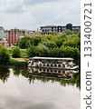 Passenger ferry moored on Vistula River reflecting on calm water surface with green trees and modern residential buildings on opposite bank on cloudy summer day in Krakow, Poland. Zablocie, Krakow 133400721