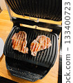 Juicy pork steaks sizzling on electric grill, positioned on wooden kitchen counter, create mouthwatering and convenient meal perfect for lunch or dinner. Pork steaks grilling on electric griddle 133400723