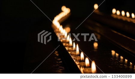 A long row of burning tea lights glows in the darkness, reflecting on a polished surface. This serene and solemn image is perfect for memorials, vigils, or spiritual themes. A long row of burning tea lights glows in the darkness, reflecting on a polished surface. This serene and solemn image is perfect for memorials, vigils, or spiritual themes. 133400899