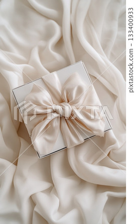 A white gift box with a beige silk bow on draped satin. This elegant, romantic scene is perfect for weddings, anniversaries, holidays, and luxury branding concepts. A beautiful surprise. 133400933