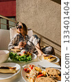 Young woman in pajamas and sunglasses enjoying breakfast on balcony, soaking up morning sun while savoring healthy meal of omelette, tomatoes, and coffee. Woman wearing pajamas relaxing on balcony 133401404