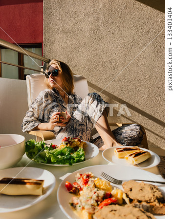 Young woman in pajamas and sunglasses enjoying breakfast on balcony, soaking up morning sun while savoring healthy meal of omelette, tomatoes, and coffee. Woman wearing pajamas relaxing on balcony Young woman in pajamas and sunglasses enjoying breakfast on balcony, soaking up morning sun while savoring healthy meal of omelette, tomatoes, and coffee. Woman wearing pajamas relaxing on balcony 133401404