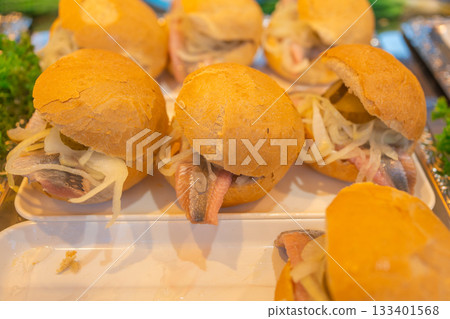 A batch of herring sandwiches arranged closely on a tray at a festival food stall. The bun texture, onions, and fish create a characteristic look of local street cuisine. 133401568