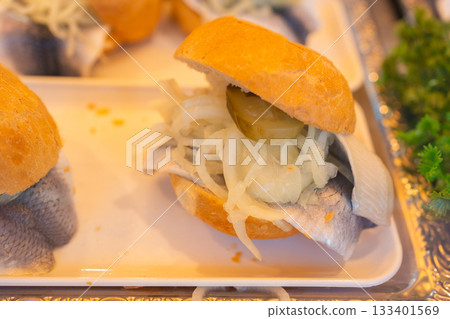 A close-up of a traditional fish sandwich with marinated herring, sliced onions, and pickles on a small bread roll. Fresh ingredients and soft lighting create an authentic street-food atmosphere A close-up of a traditional fish sandwich with marinated herring, sliced onions, and pickles on a small bread roll. Fresh ingredients and soft lighting create an authentic street-food atmosphere 133401569