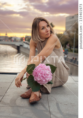 A woman sits by the Moscow riverbank at sunset, holding a large pink hydrangea against warm pastel sky tones and calm water reflections 133401973