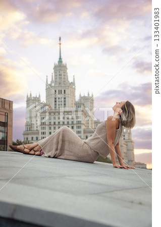 A woman sits on a rooftop at sunset with the Kotelnicheskaya Embankment Building rising behind her near Tessinsky Bridge, surrounded by pastel sky 133401983