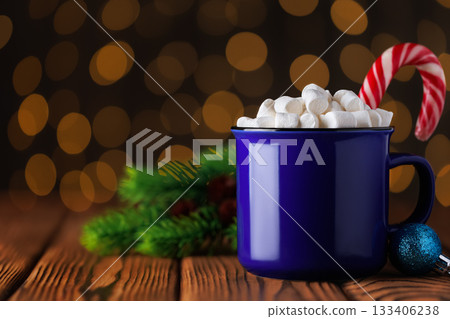 Blue cup of hot cocoa with marshmallows and candy cane on wooden table against background of bokeh glowing garland lights 133406238
