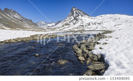 Scenic winter view of Leirvassbu with snow-covered mountains and a flowing river in Jotunheimen National Park 133409095