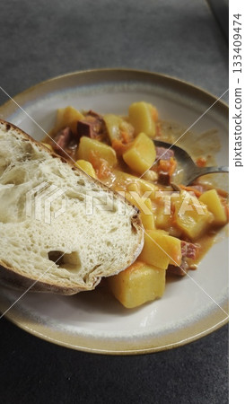 Stew with potatoes, carrots and sausage served with a slice of fresh bread on a ceramic plate 133409474