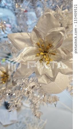 A shimmering silver poinsettia ornament with golden details decorates a bright white Christmas tree. Symbolizes festive elegance, winter beauty and the sparkle of holiday celebration 133409521