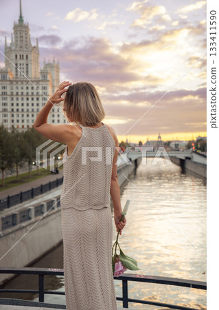 A woman stands on the Tessinsky Bridge at sunset, holding a pink flower stem while overlooking the river and the Kotelnicheskaya Embankment Building A woman stands on the Tessinsky Bridge at sunset, holding a pink flower stem while overlooking the river and the Kotelnicheskaya Embankment Building 133411590