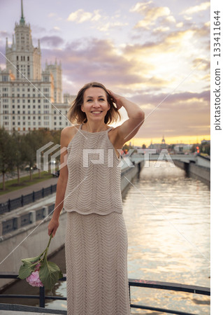 A woman stands on the Tessinsky Bridge at sunset, holding a pink flower stem with the Kotelnicheskaya Embankment Building glowing in the background A woman stands on the Tessinsky Bridge at sunset, holding a pink flower stem with the Kotelnicheskaya Embankment Building glowing in the background 133411644