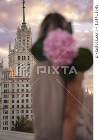 A woman holds a pink hydrangea in the foreground, with the Kotelnicheskaya Embankment Building rising sharply in the background under a sunset sky A woman holds a pink hydrangea in the foreground, with the Kotelnicheskaya Embankment Building rising sharply in the background under a sunset sky 133411645