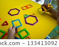 A kid explores spatial relationships by playing with a magnetic constructor featuring colorful geometric shapes, creating three-dimensional forms. 133413092