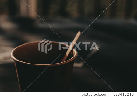 Steaming coffee in a paper cup with a wooden stirrer, close-up shot. A warm cup of takeaway coffee sits on a table Steaming coffee in a paper cup with a wooden stirrer, close-up shot. A warm cup of takeaway coffee sits on a table 133413216