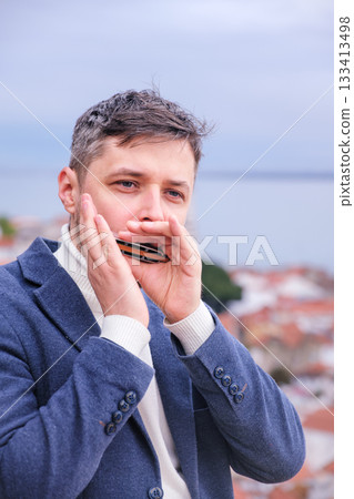 Man musician in a blue blazer and white sweater playing blues on a harmonica with eyes closed, standing outdoors in city street Man musician in a blue blazer and white sweater playing blues on a harmonica with eyes closed, standing outdoors in city street 133413498