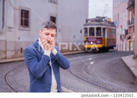 Man musician in a blue blazer and white sweater playing blues on a harmonica with eyes closed, standing outdoors in Lisbon city street, Portugal with tram in background 133413501