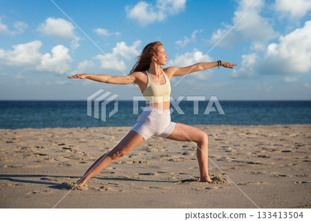 A woman performs the Virabhadrasana 2 Warrior 2 pose on a sandy beach, backlit by a beautiful sunset, enhancing her peaceful yoga practice. 133413504